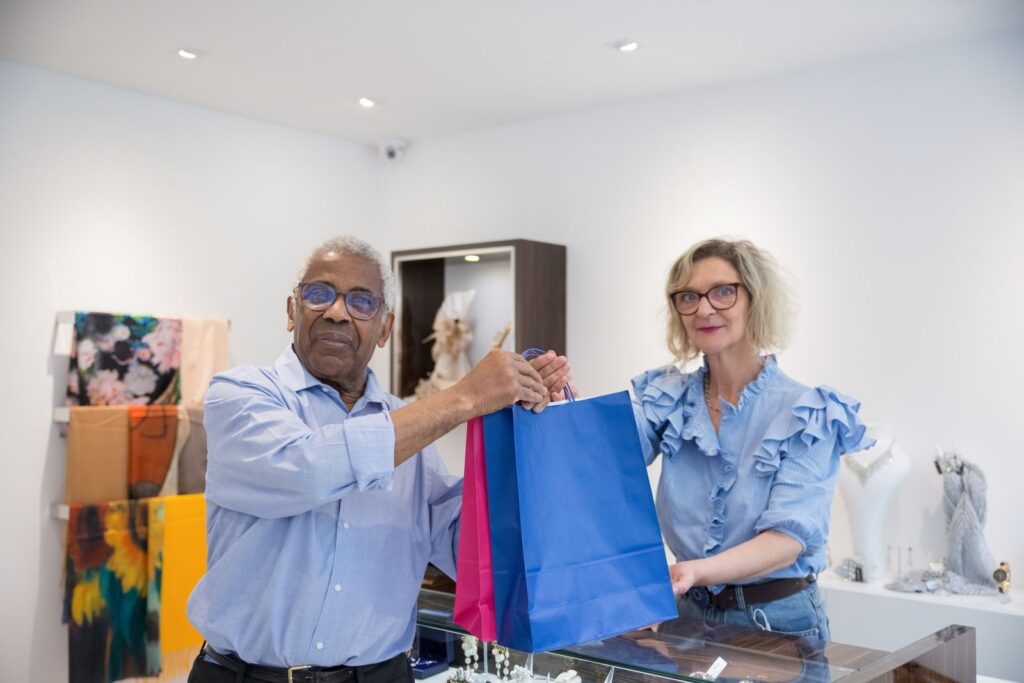 An elderly man and woman shopping together in a clothing store, carrying colorful bags.