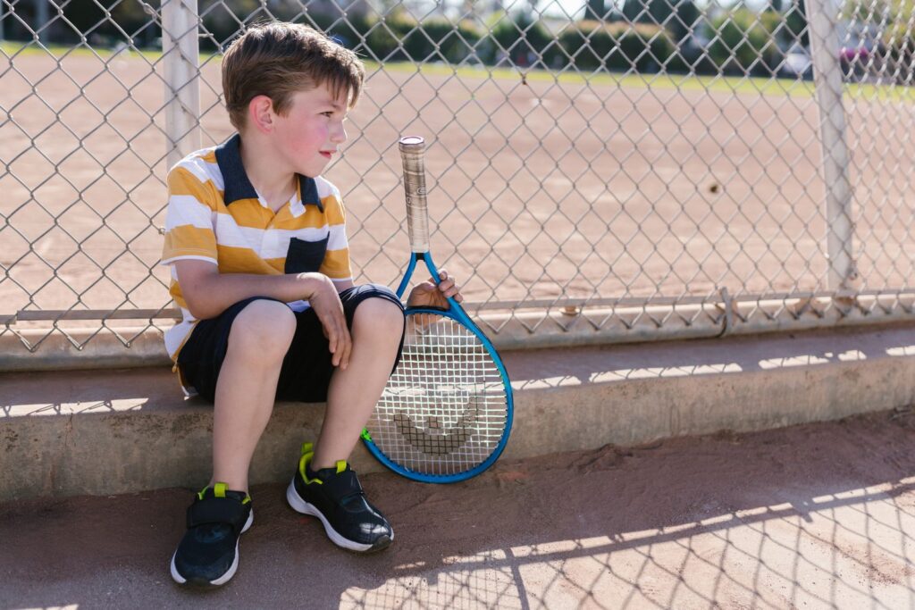 A young boy sits with a tennis racket by a chain-link fence on a sunny day, embodying youthful leisure.