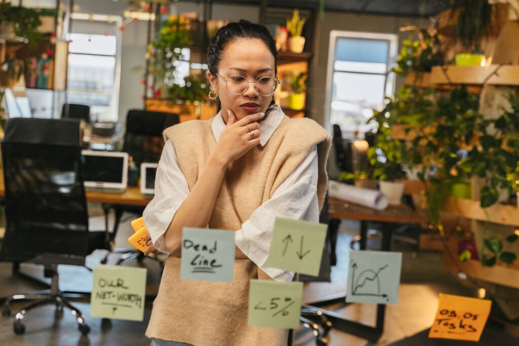 Asian woman thinking in green office with sticky notes on glass, workplace planning concept.