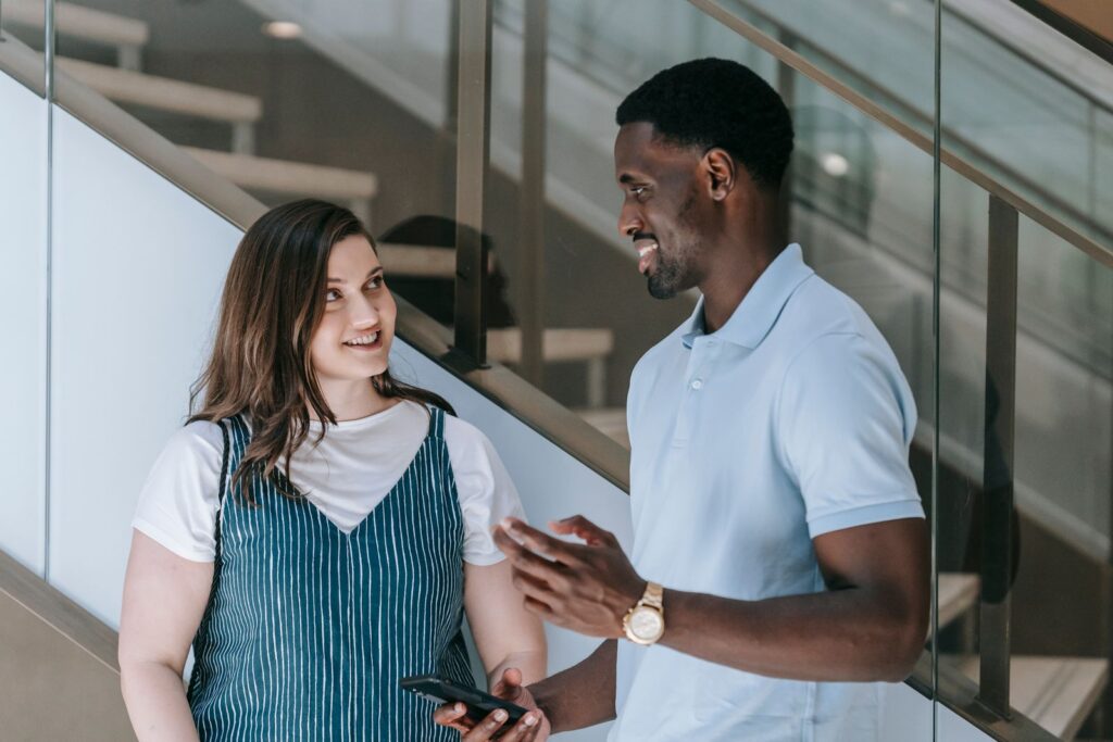 A man and a woman smiling and talking indoors by a staircase.