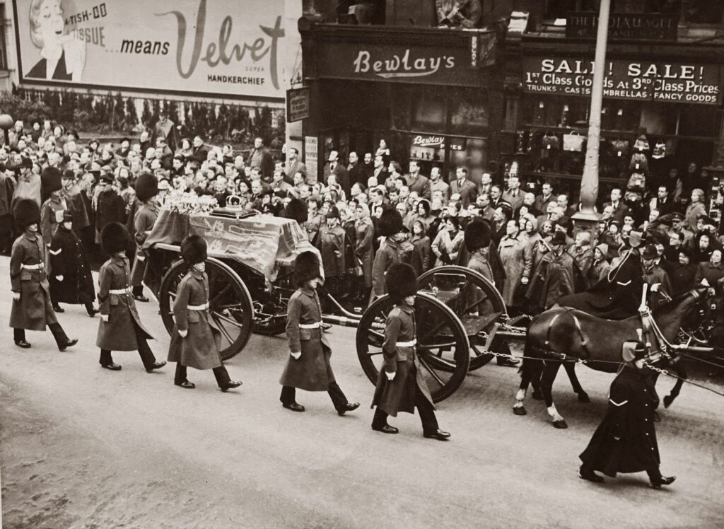 Historical black and white photo of a royal funeral procession with soldiers and crowd in city street.
