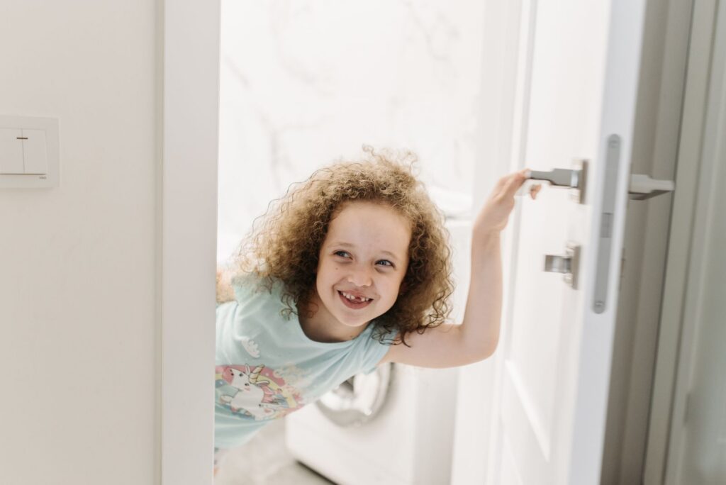 Joyful child with curly hair smiling as she peeks through a doorway indoors.