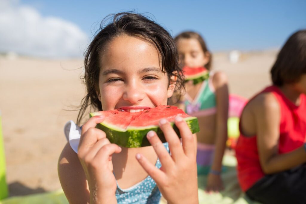 Smiling children enjoying watermelon slices at a sunny beach, capturing summer bliss.