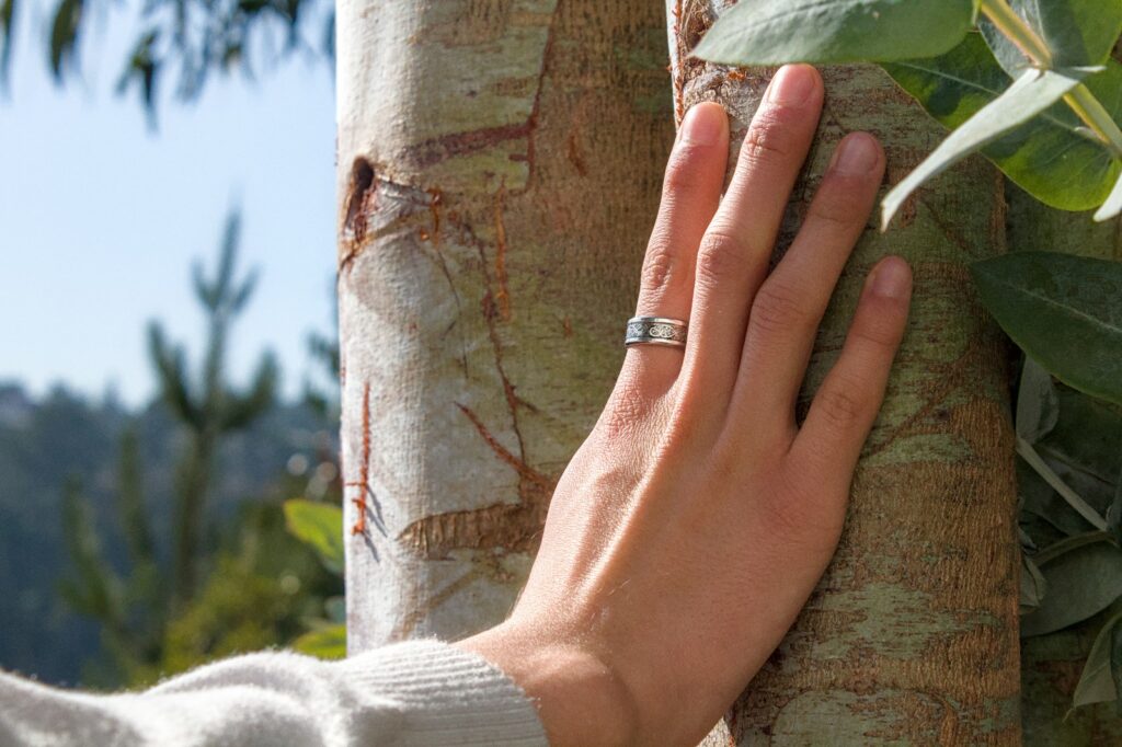 A hand with a ring gently touching a tree trunk.
