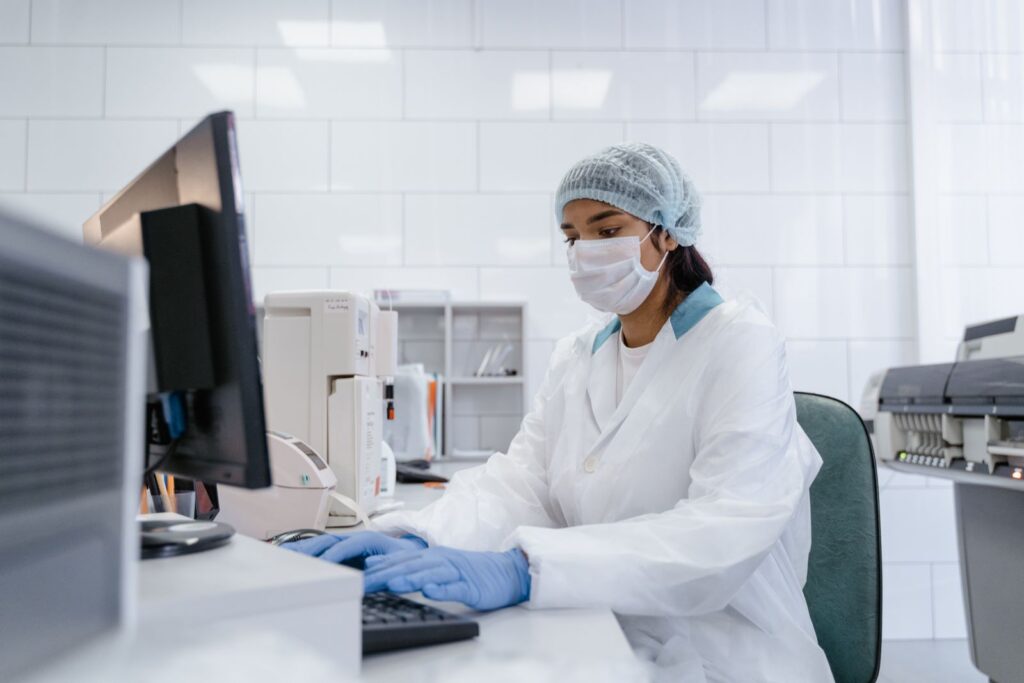Scientist using a computer in a laboratory with protective gear including gloves, mask, and hair net.