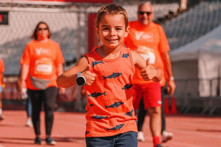 Young boy in orange tank top with fish design smiling and giving thumbs up during outdoor event.