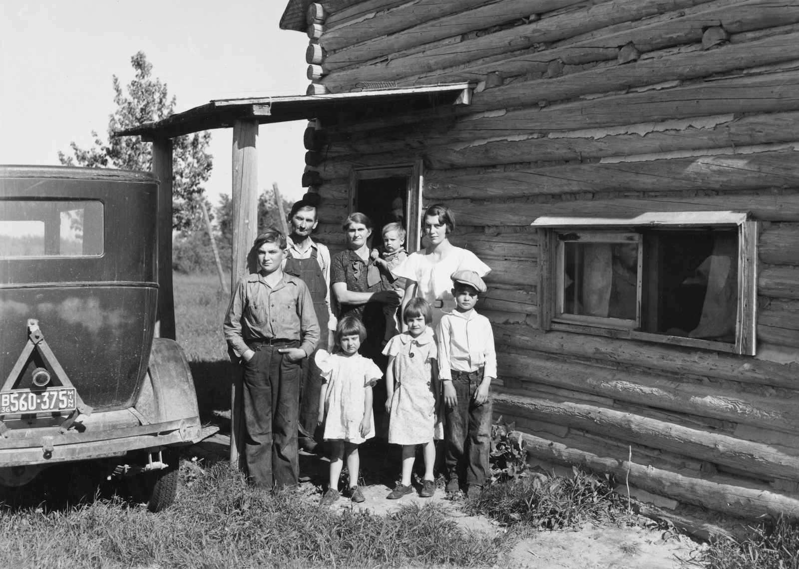Family poses outside a rustic log cabin with a car.